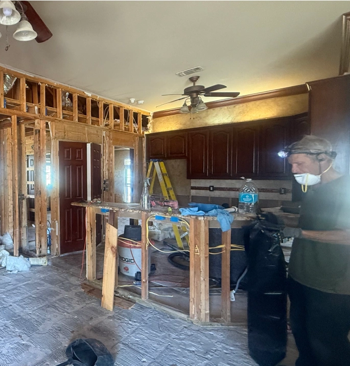 A man in a respirator mask and headlamp demolishes the interior of a house, revealing wooden wall studs.